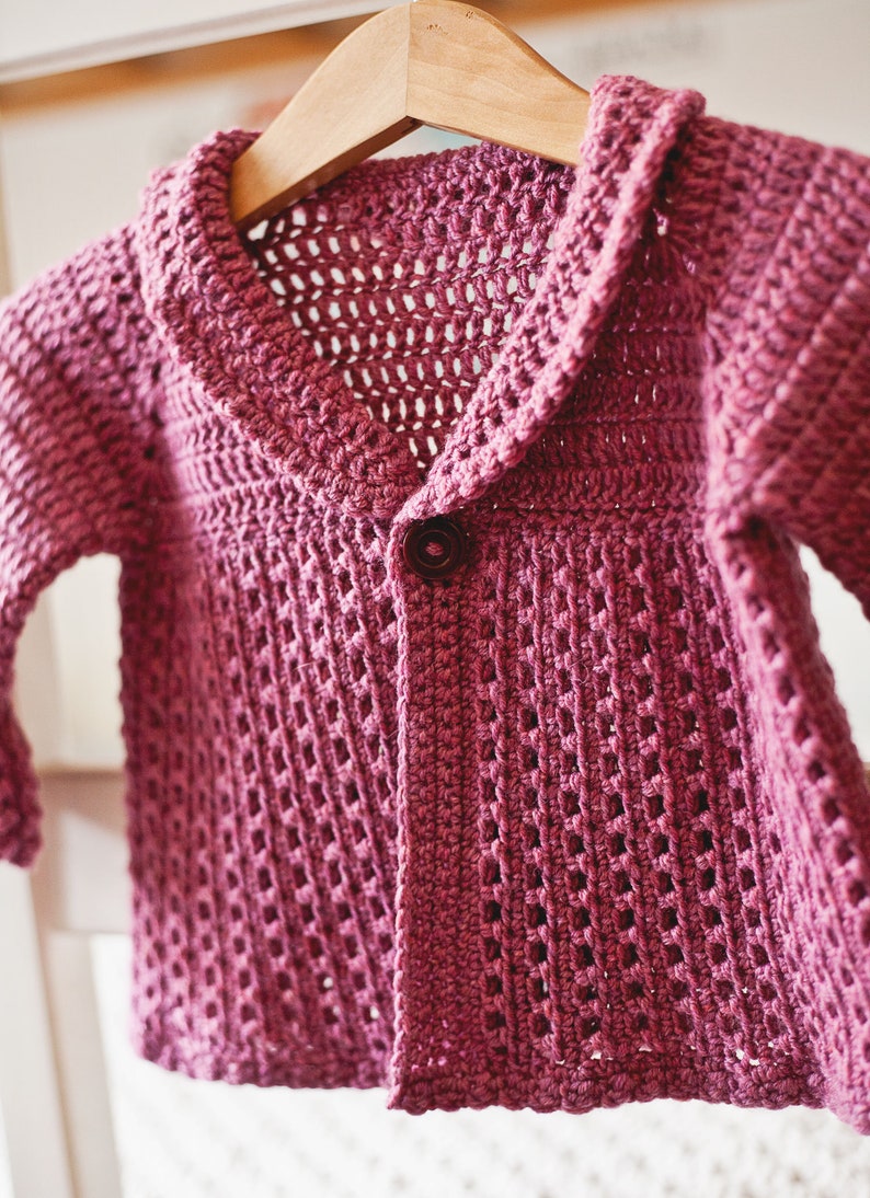 May include: A pink crocheted cardigan with a button closure and a wide collar. The cardigan is hanging on a wooden hanger.