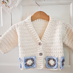 Puede incluir: Rebeca de bebé de crochet color crema, con cuello en V y mangas largas. El suéter presenta dos bolsillos cuadrados con detalles azules y marrones y se abrocha con dos botones decorativos. La rebeca está colgada de una percha de madera.