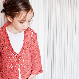 May include: A coral-colored, crocheted cardigan with a scalloped edge and a single red button. The openwork design of the sweater is visible, and it is worn over a white shirt. The cardigan has long sleeves and is worn by a young person.