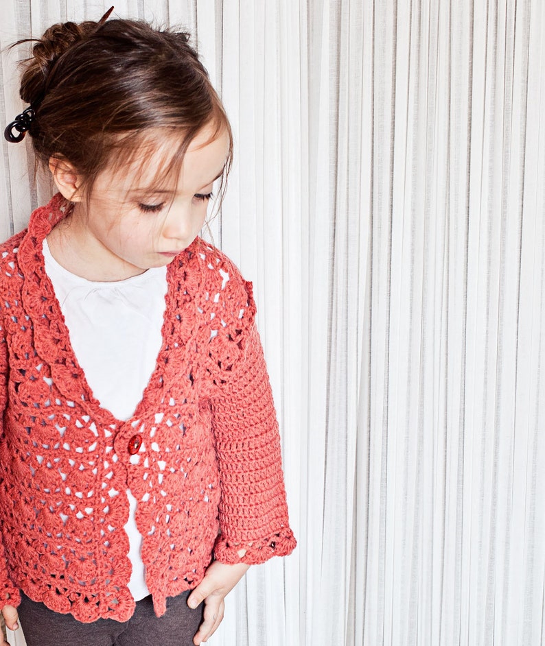 May include: A coral-colored, crocheted cardigan with a scalloped edge and a single red button. The openwork design of the sweater is visible, and it is worn over a white shirt. The cardigan has long sleeves and is worn by a young person.