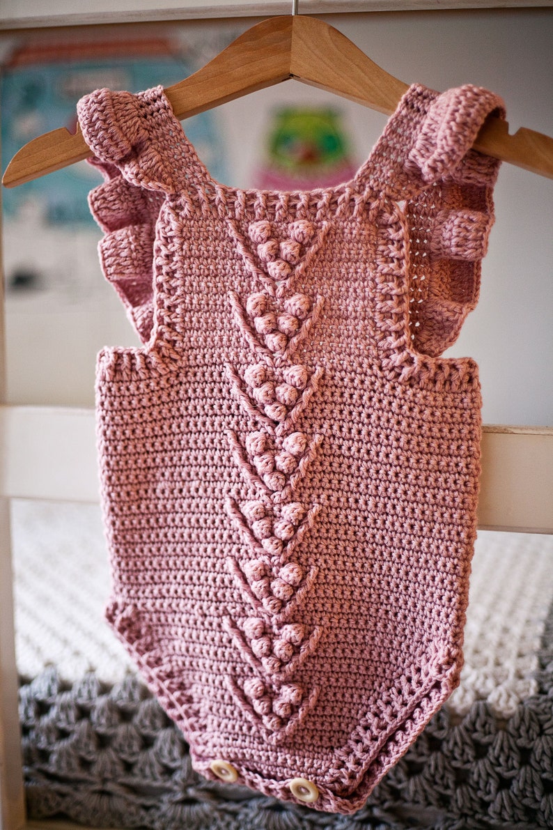 May include: A pink crocheted baby onesie with a ruffled neckline and a decorative stitch pattern. The onesie has a button closure at the bottom.