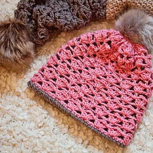 Op de afbeelding: Een roze gebreide beanie met een bruine nep bont pompon. De beanie is gemaakt met een delicaat open steekpatroon.