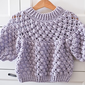 May include: A lilac-coloured, hand-crocheted sweater with a textured, bubble-like stitch pattern. The sweater has a round neckline and puffy, three-quarter length sleeves. It is displayed on a wooden hanger against a white background.