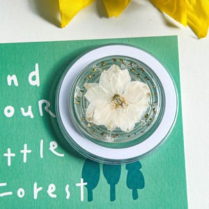Puede incluir: Un soporte de teléfono redondo y transparente con una flor blanca y motas doradas encapsuladas en resina, sobre un fondo blanco y verde. La tarjeta verde tiene el texto "Find your little Forest" en blanco. Una flor amarilla en el fondo.