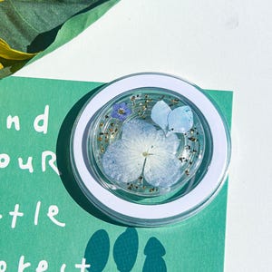 May include: A clear, round phone grip with a white border, containing a blue flower and gold glitter, rests on a green card. The card has the text "Find your little Forest" and tree silhouettes. A yellow sunflower is in the background.