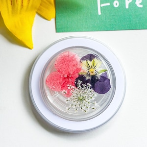 May include: A clear resin coaster with pressed flowers in pink, purple, yellow, and white. The coaster has a white border. A green card with the word "Forest" is visible. The coaster is set against a white background.