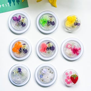 May include: Nine clear, round resin coasters with pressed flowers and a strawberry. The coasters feature a variety of colourful flowers, including purple, orange, pink, and yellow, set in clear resin within a white border. The strawberry coaster is red with a green stem.