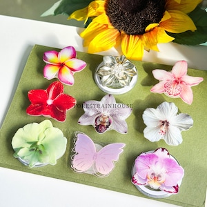 May include: A collection of floral phone grips in various colors, including pink, red, white, and purple. The grips feature flower and butterfly designs, with a sunflower in the background. The text "FORESTRAINHOUSE" is visible.