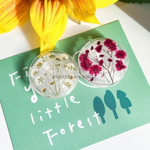 Könnte beinhalten: Zwei klare, runde Harz-Untersetzer mit getrockneten Blumen. Einer hat weiße Blüten, der andere leuchtend rote. Die Untersetzer liegen auf einer türkisfarbenen Karte mit dem Text "tiny little Forest". Eine gelbe Blume ist im Hintergrund.