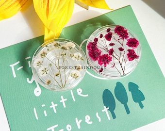 May include: Two clear, round resin coasters with dried flowers. One coaster features white flowers, the other has vibrant red flowers. The coasters rest on a teal card with the text "tiny little Forest". A yellow flower is in the background.