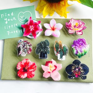 Puede incluir: Una colección de pines acrílicos con temática floral y vegetal. Los pines presentan varios diseños de flores y plantas, incluyendo un lirio rojo, plumeria rosa y una flor púrpura. El texto "Find your little Forest" es visible en una tarjeta verde.