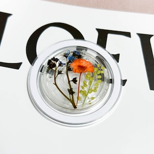 May include: A clear, round resin coaster with a white border, containing dried flowers in orange, blue, and green. The coaster is set against a white background with black lettering.