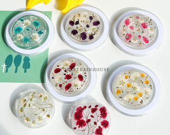 May include: A collection of clear, round phone grips with embedded dried flowers in various colors, including blue, purple, red, pink, and yellow. Each grip is set in a white circular frame. The text "FORESTRAINHOUSE" is visible on one of the grips.