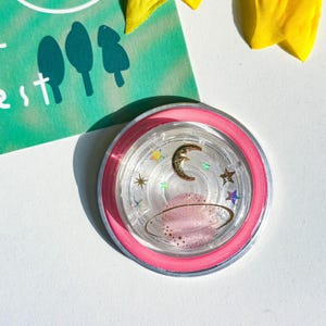 May include: A clear, round shaker with a pink rim and silver base, filled with a gold crescent moon, stars, and a pink planet. The shaker is on a white surface with a green card and yellow flower.