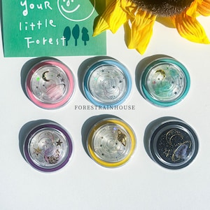 Puede incluir: Seis posavasos redondos de resina transparente con diseños celestiales, incluyendo lunas, estrellas y planetas. Cada posavasos tiene un borde de color: rosa, azul, turquesa, morado, amarillo y negro. El texto "FORESTRAINHOUSE" es visible.