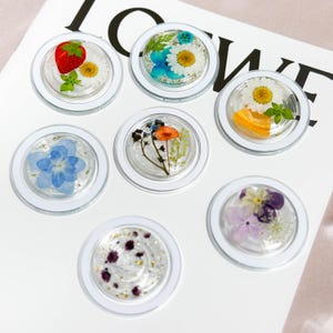 May include: Seven clear round resin paperweights with white rims, each containing a different pressed flower arrangement. The flowers include a strawberry, daisies, blue hydrangeas, orange blossoms, and purple flowers.