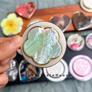 May include: A circular phone grip featuring a flower-shaped inlay made of iridescent, light green mother-of-pearl with a thin gold-toned metal border, set against a matte silver-toned base, with the text ForestHouse visible in the background.