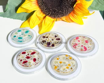 May include: Five clear, round phone grips with dried flowers and gold flakes encased in resin. Each grip has a different color scheme, including red, pink, yellow, blue, and purple flowers. The grips are set against a white background with a sunflower.