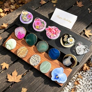 May include: A collection of circular and heart-shaped phone grips featuring various designs including pressed flowers, agate slices, and stone textures. A white card with the gold text Happy Birthday is included with the assorted phone accessories.