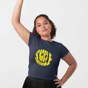 May include: Navy blue t-shirt with a yellow smiley face design. The word "SMILE" is integrated into the smiley face. The shirt is a crew neck and short-sleeved.