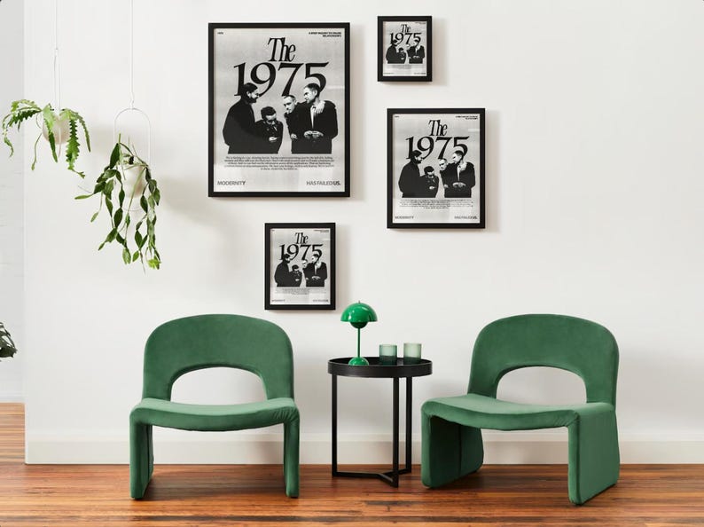 May include: Four black framed posters with the text "The 1975" in white. The posters are hanging on a white wall. Two green velvet chairs and a black side table with a green lamp are in front of the wall.