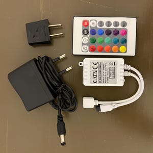 May include: A white LED light strip controller with a remote control, power adapter, and a power supply. The remote control has 16 buttons for different colors and modes. The power adapter is black and has a plug. The power supply is white and has a label that says "CATA CE CT-946 RGB KUMANDA".