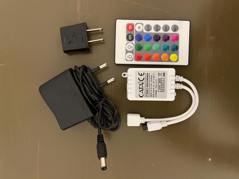 May include: A white LED light strip controller with a remote control, power adapter, and a power supply. The remote control has 16 buttons for different colors and modes. The power adapter is black and has a plug. The power supply is white and has a label that says "CATA CE CT-946 RGB KUMANDA".