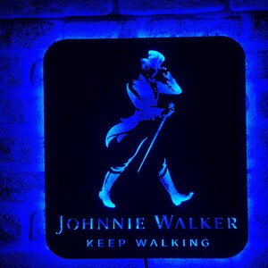 May include: A black and blue illuminated sign with the Johnnie Walker logo. The logo features a man in a walking pose with a cane. The text "Keep Walking" is below the logo.