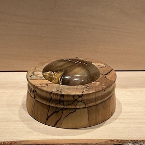 Peut inclure: Une boîte ronde en bois avec un couvercle. Le couvercle présente une pierre polie de forme ovale dans les tons marron et or. La boîte et le couvercle sont en bois clair avec des motifs de grain sombres et complexes. La boîte repose sur une surface en bois clair.