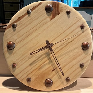 May include: A wooden clock face with a light brown wood grain and copper-colored accents. The clock hands are black metal and point to the time.
