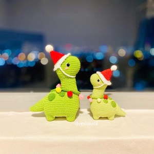 May include: Two green crocheted dinosaur toys wearing red Santa hats with white trim. The larger dinosaur has a white string of lights around its neck. The smaller dinosaur is standing behind the larger dinosaur. The text "demiwool" is visible in the bottom center of the image.