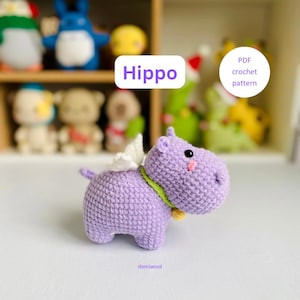 May include: A purple crocheted hippopotamus with white wings and a green collar. The hippo is standing on a white surface. The text "Hippo" is written in purple above the hippo. The text "PDF crochet pattern" is written in white on a purple circle in the upper right corner of the image.