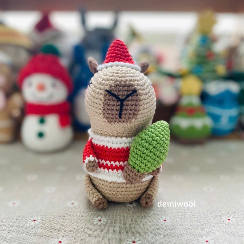 May include: A crocheted llama wearing a red and white striped sweater and a red Santa hat, holding a green Christmas tree. The llama is tan with brown accents. Other crocheted holiday figures are in the background.
