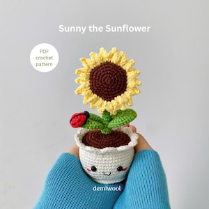 Könnte beinhalten: Eine gehäkelte Sonnenblume mit brauner Mitte und gelben Blütenblättern in einem weißen Topf mit einem lächelnden Gesicht. Ein roter Marienkäfer sitzt auf dem Stiel der Sonnenblume. Der Text "PDF crochet pattern" und "Sunny the Sunflower" ist auf dem Bild zu sehen.