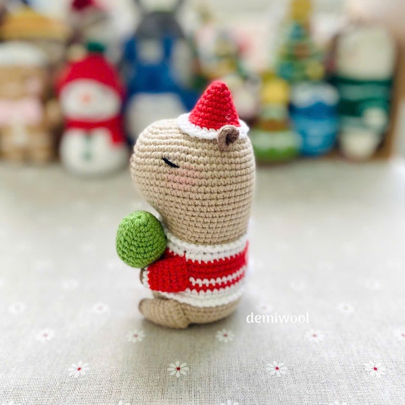 May include: A crocheted capybara figure wearing a red and white striped sweater and a red Santa hat. The capybara is holding a green ball. The figurine has closed eyes and a smiling expression. The text "demiwool" is visible on the right side.