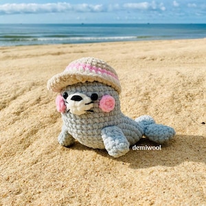 May include: A grey crocheted seal wearing a pink and white striped sun hat. The seal is sitting on a sandy beach.