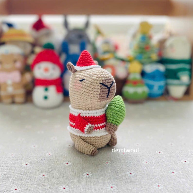 May include: A crocheted capybara wearing a Santa hat and a red and white sweater, holding a small green Christmas tree. Other crocheted holiday figures are in the background. The word "demiwool" is in the lower right corner.