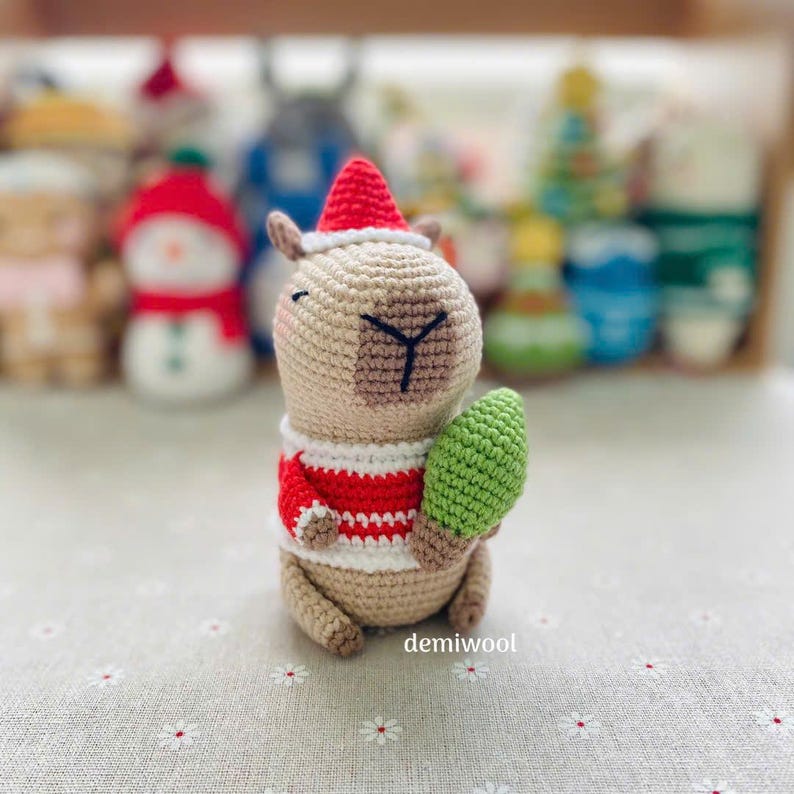 May include: A crocheted capybara wearing a Santa hat and a red and white striped sweater, holding a green Christmas tree. The handmade plush toy has a beige body, brown face patch, and black embroidered details. The word "demiwool" is visible.