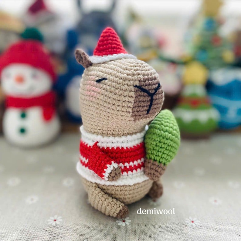 May include: A crocheted capybara wearing a red and white striped sweater and a Santa hat, holding a green Christmas tree. The handmade plush toy is beige with brown accents and black facial details. Other holiday-themed crochet figures are in the background.