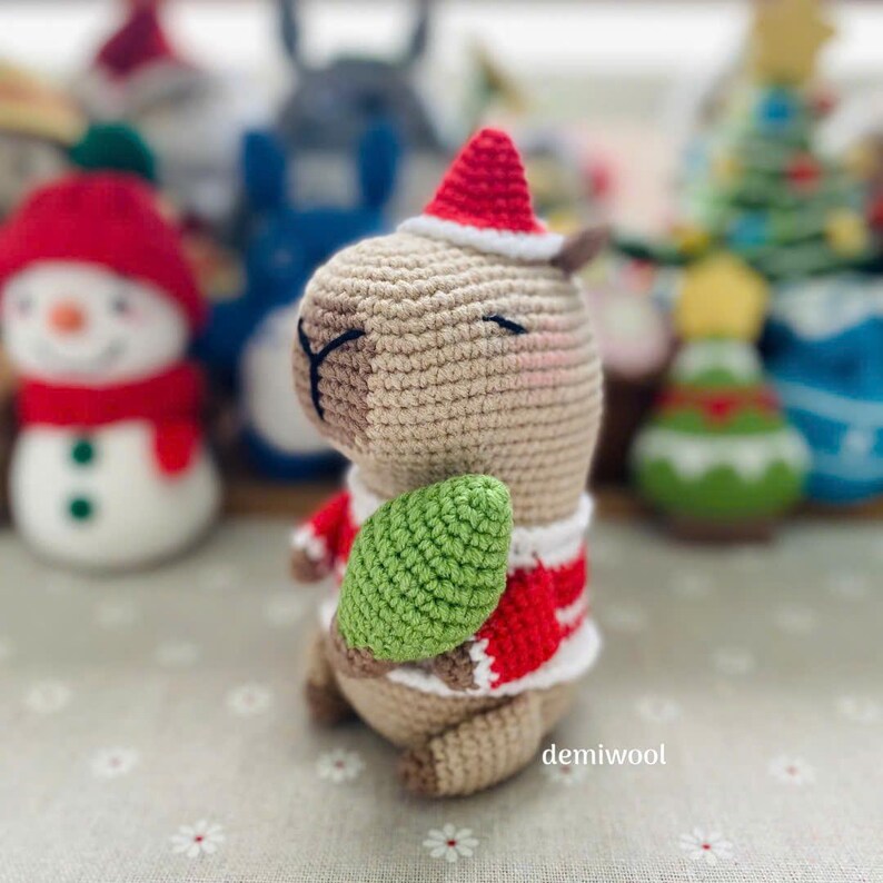 May include: A crocheted Christmas capybara wearing a red and white Santa hat and sweater, holding a green leaf. Other crocheted holiday figures are in the background, including a snowman and Christmas trees. The word "demiwool" is visible.