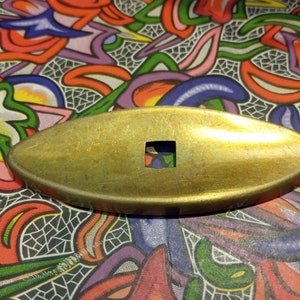 May include: A gold-colored metal oval-shaped object with a square hole in the center. The object is lying on a colorful patterned surface.