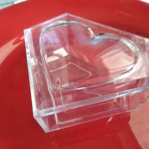 May include: A clear plastic box shaped like a house with a heart-shaped opening in the front. The box is sitting on a red surface.