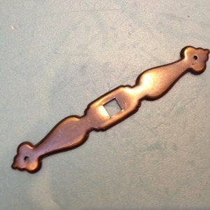 May include: A long, thin, antique-looking, copper-colored metal door handle with a square hole in the center and decorative details on the ends.