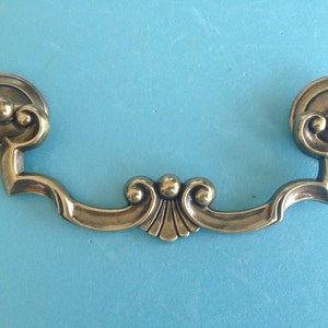 May include: A vintage-style brass drawer pull with a decorative design. The pull features two circular end pieces and a curved handle with ornate detailing. The brass finish has a warm, antique appearance, suitable for furniture restoration or decorative projects.