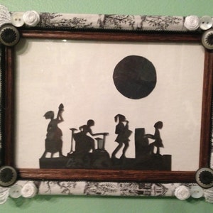 May include: A black and white silhouette of five figures in a framed picture with a black and white patterned border. The figures are silhouetted against a white background with a black circle in the upper right corner.