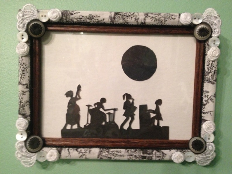 May include: A black and white silhouette of five figures in a framed picture with a black and white patterned border. The figures are silhouetted against a white background with a black circle in the upper right corner.