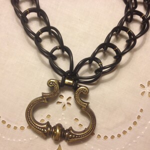 May include: A black rubber chain necklace with a gold-toned ornate pendant. The pendant is shaped like a heart with a decorative design.