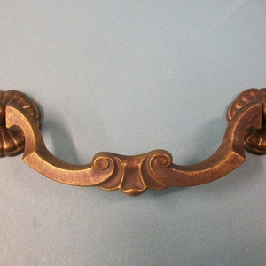 May include: A brass cabinet pull with a decorative, ornate design. The pull has a curved shape with a center section that features a scroll-like pattern. The ends of the pull are rounded and have a floral design.