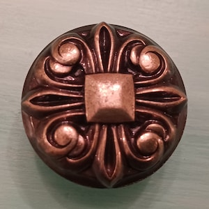 May include: A decorative, ornate, bronze-colored metal knob with a square center and intricate floral designs.
