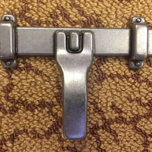May include: A silver-toned metal latch with a T-shaped handle. The latch has a rectangular base with two decorative ends and a raised center section. The handle is attached to the center section and has a U-shaped opening.